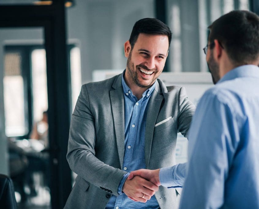 In an office setting, two men from the Asset Valuations Group are shaking hands and smiling. One is wearing a gray suit jacket and blue shirt, while the other dons a light blue shirt. The backdrop reveals large windows and blurred office furniture, creating a professional ambiance.