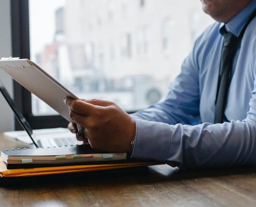 A person in a blue shirt and tie sits at a desk, clipboard in hand. Surrounded by a laptop, notebook, and documents from the Asset Valuations Group, they appear focused in an office setting with large windows framing the background.