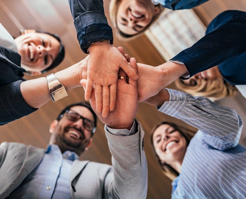 A group of five people, part of the Asset Valuations Group, stand in a circle viewed from below. They are smiling and stacking their hands together in the center, showcasing teamwork and unity against a backdrop of wooden-paneled ceilings.