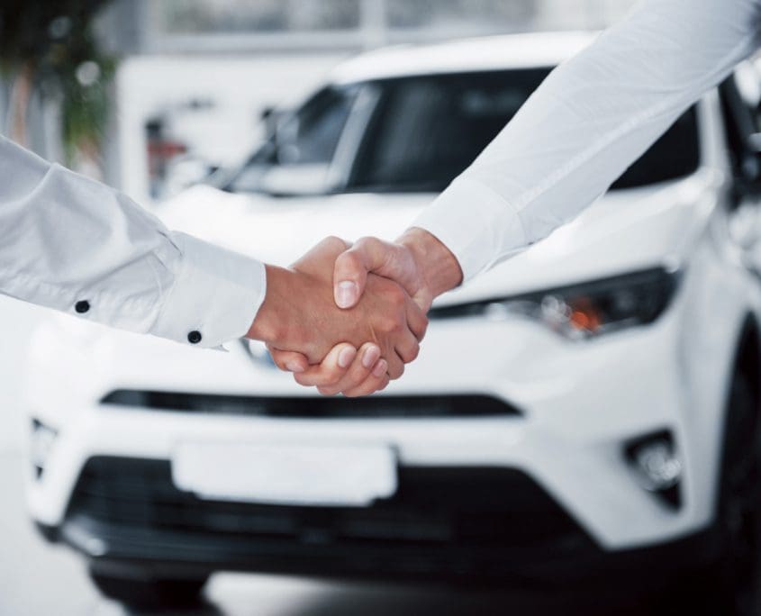 Two people, members of the Asset Valuations Group, are shaking hands in the foreground, both wearing crisp white shirts. In the background, a white car signifies a successful transaction at a dealership.