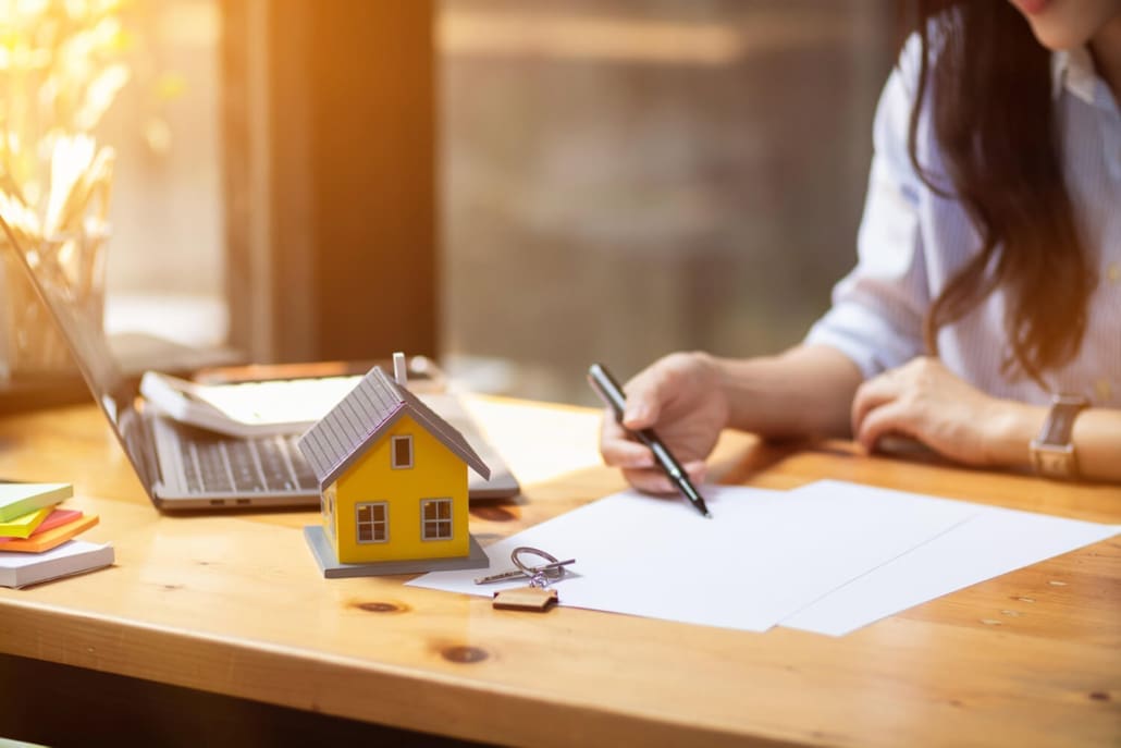 Property Valuers Brisbane, A person sits at a desk with a small model house, keys, and documents, writing on paper—perhaps consulting Property Valuers Brisbane. A laptop and colorful notepads are also on the wooden desk, with sunlight streaming in through a window.