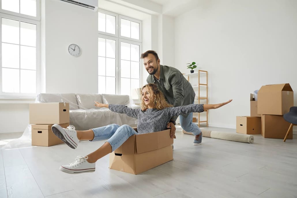Property-valuers-valuations-sydney A smiling man pushes a laughing woman who is sitting in a cardboard box with her arms outstretched. Surrounded by moving boxes in a bright, unfurnished Sydney room, they share a joyful moment during their move.