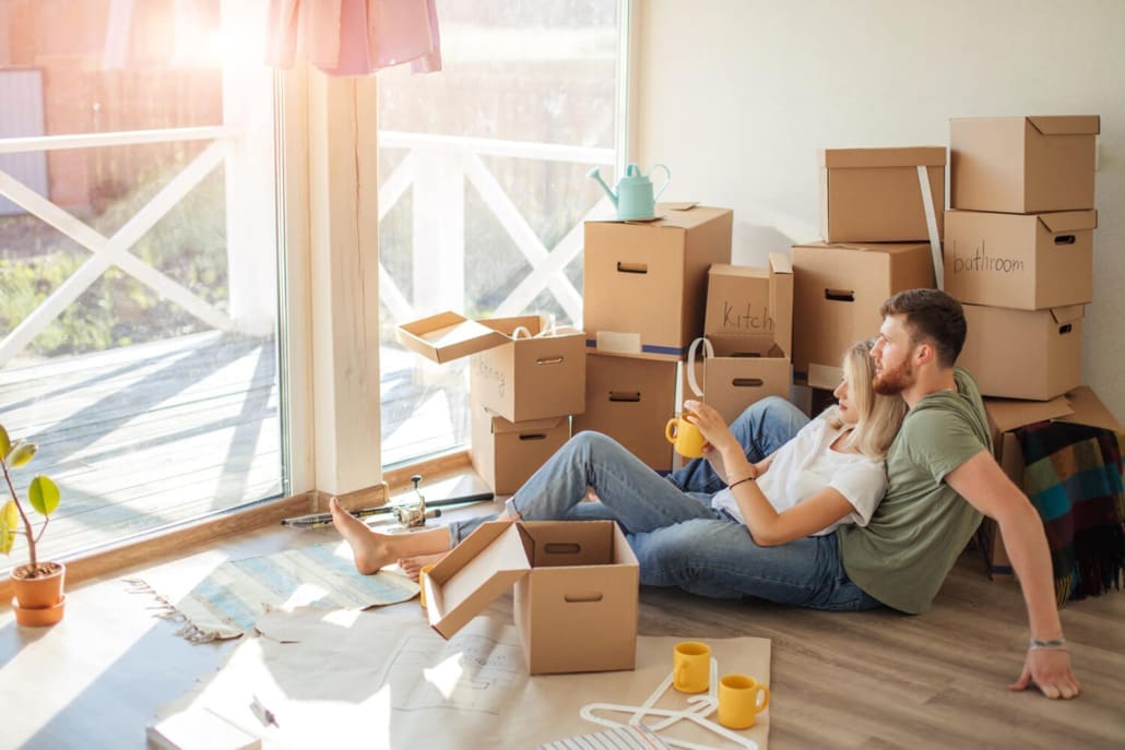 House Valuations Rockhampton, A couple sits on the floor of a sunlit room, surrounded by moving boxes and household items. Relaxing with mugs in hand, they look out the window, savoring their new beginning after securing the home through Property Valuations Rockhampton.