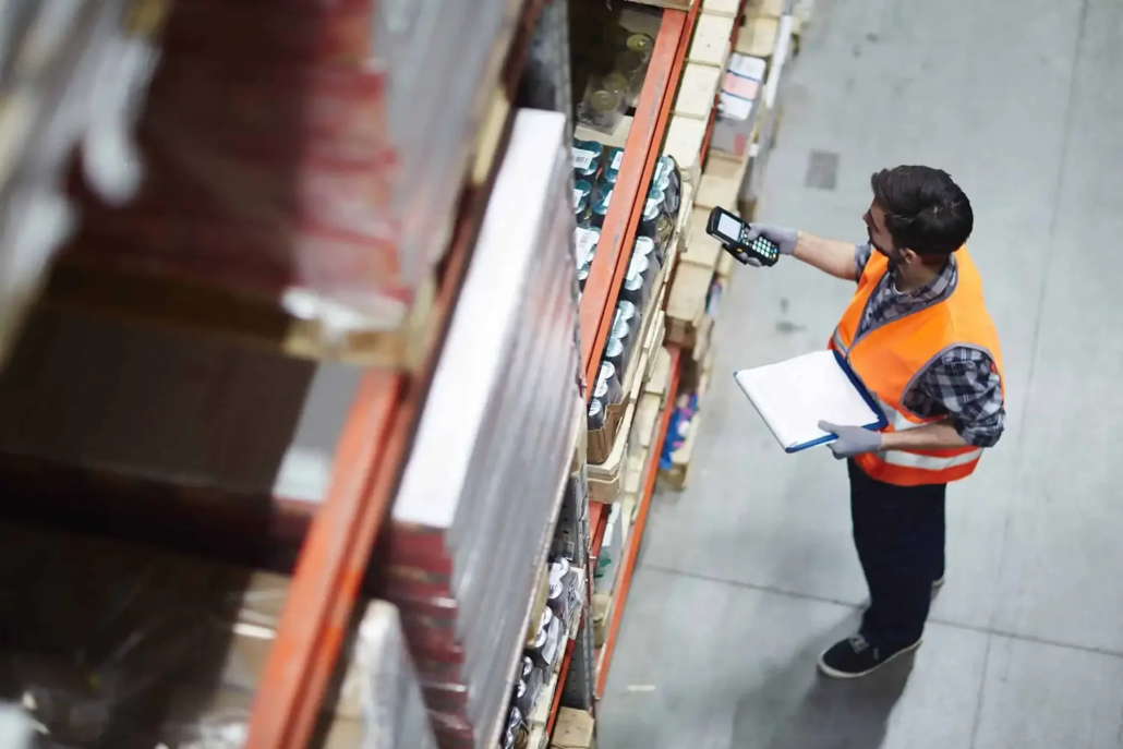 A warehouse worker wearing an orange safety vest scans items on shelves with a handheld device while holding a clipboard, conducting a stocktake in a Brisbane facility aisle lined with neatly stacked inventory.