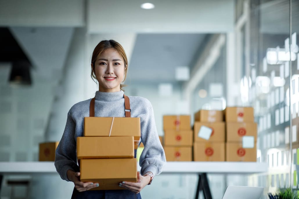 Stocktake Sydney Stocktake Sydney - A woman wearing a gray sweater and apron smiles while holding three stacked cardboard boxes. In the background, more boxes rest on a white table in a bright, modern office space during a Stocktake Sydney event.