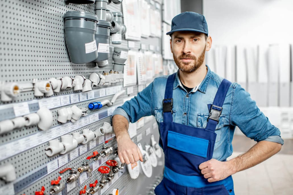 Superannuation Valuations, A man wearing a blue cap and overalls stands in a hardware store, leaning on a display wall filled with plumbing pipes, fittings, and valves—perhaps contemplating Superannuation Valuations as he plans his future.
