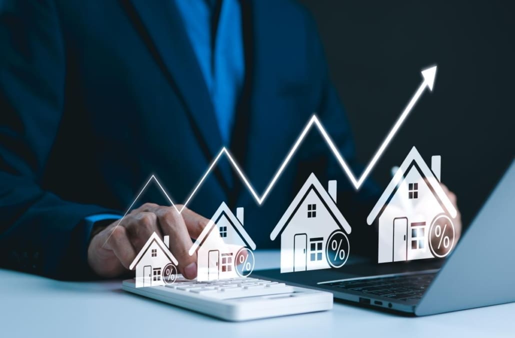A person in a suit uses a calculator and laptop, with illustrated houses, percentage signs, and an upward trending graph overlayed, symbolizing rising real estate or mortgage rates and highlighting the impact of Valuation Turnaround Times.