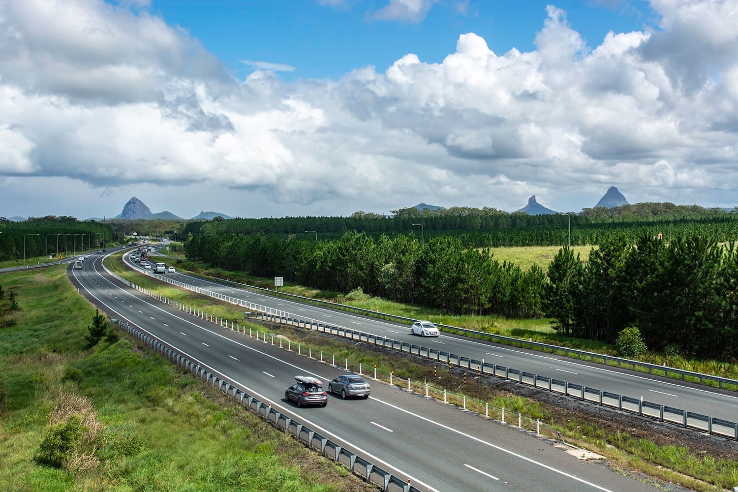 The Bruce Highway in the early morning making its way south to B A divided highway with cars traveling in both directions near Valuations Rockhampton, surrounded by green trees and fields under a partly cloudy sky, with distant mountains visible on the horizon.