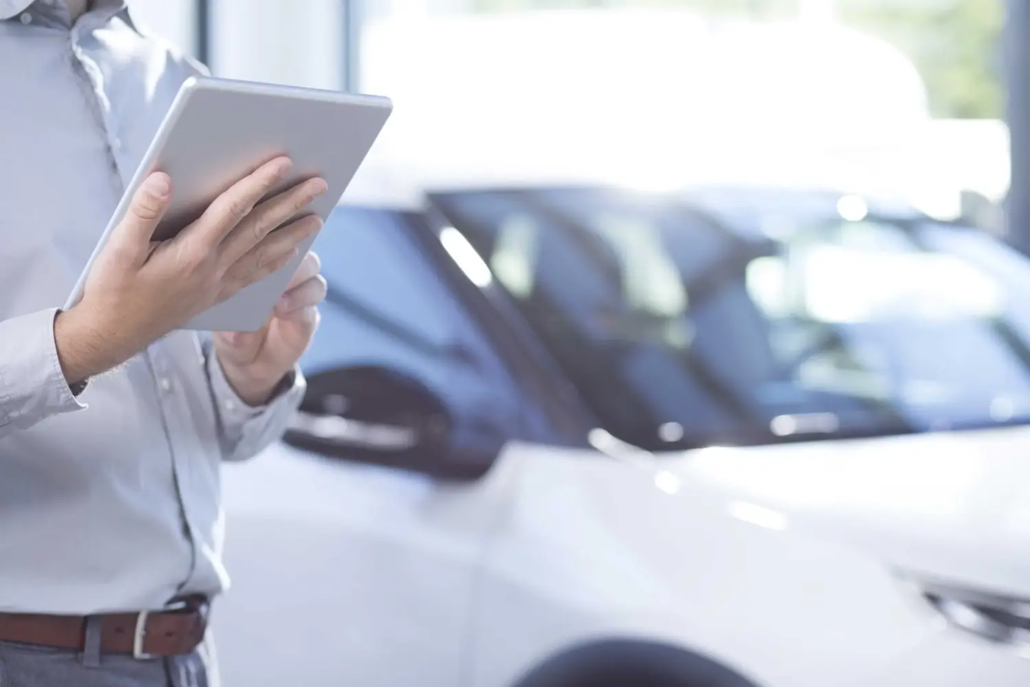 valuers-valuations-sydney A person holding and using a tablet stands next to a white car in a bright indoor Sydney showroom or dealership. The person's face is not visible.