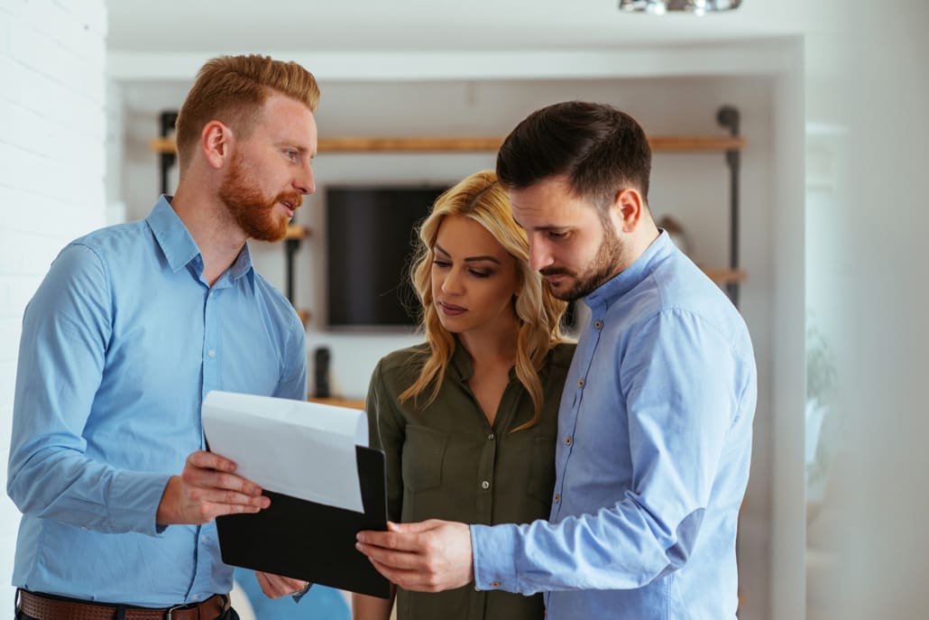 why-do-i-need-a-property-valuation A man with a clipboard shows documents to a couple, who are closely reviewing the papers together in a bright, modern interior setting, discussing questions like "Why Do I Need a Property Valuation?.