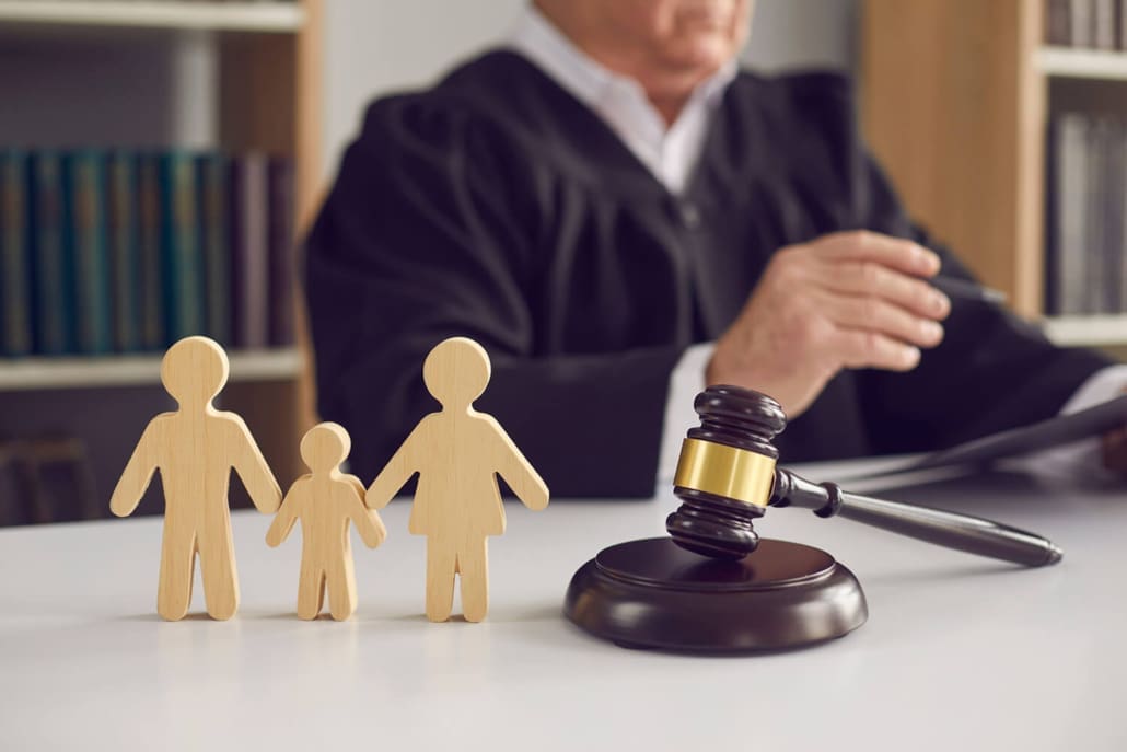 Compliant With Family Court Requirements - A judge sits at a desk with a gavel, holding documents. In the foreground are wooden cutouts of two adults and a child, symbolizing a family, suggesting a legal decision involving family law.