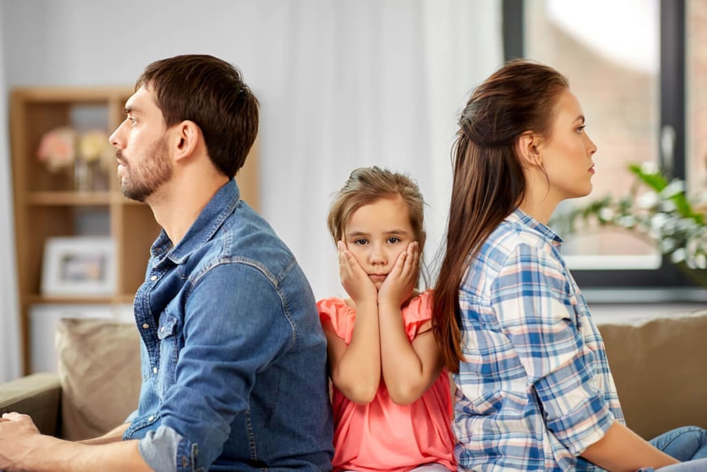 When Are Family Law Valuations Required - A man and woman sit back-to-back on a couch, looking away from each other. A young girl sits between them, holding her face in her hands and looking concerned. The scene suggests family tension.