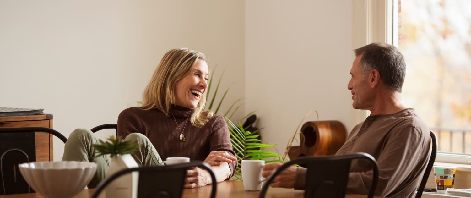 Superannuation Valuations, Two adults sit at a wooden dining table, smiling and laughing together over coffee. As sunlight streams in, they discuss superannuation valuations in a warm, cozy atmosphere. A plant and kitchen items add to the inviting scene.