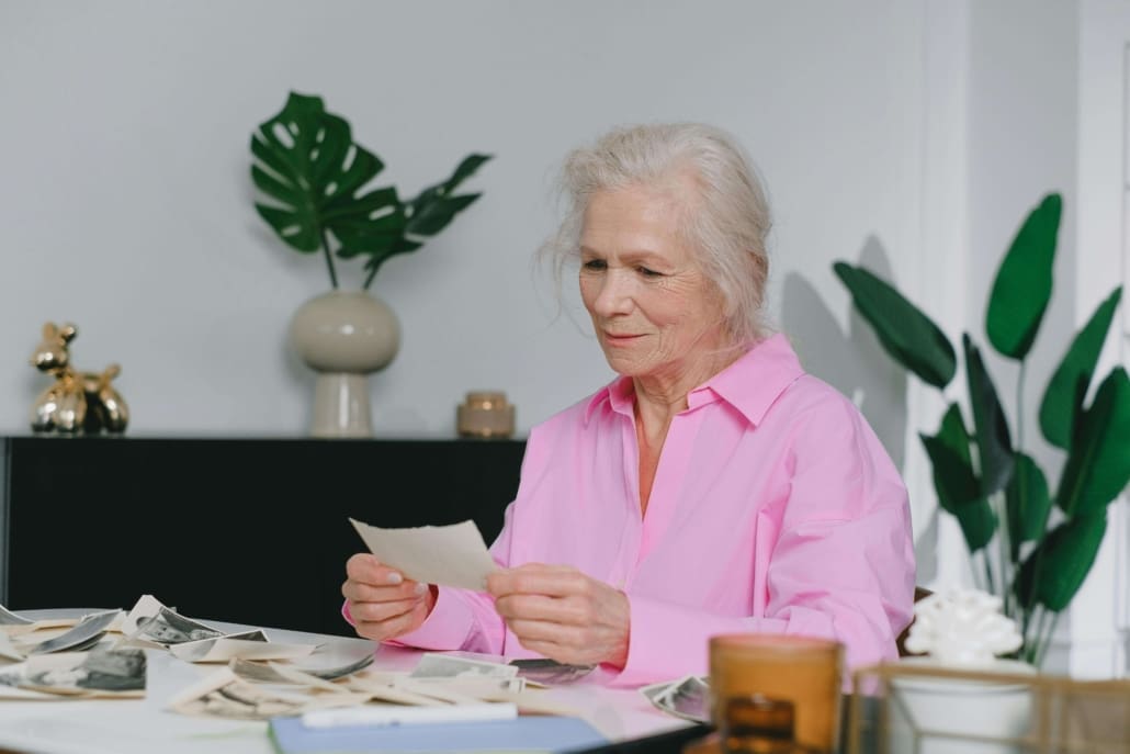 An elderly woman with gray hair wearing a pink shirt sits at a table, looking at old photographs related to Deceased Estate CGT Valuations. The table is scattered with more photos, and green plants are in the background.