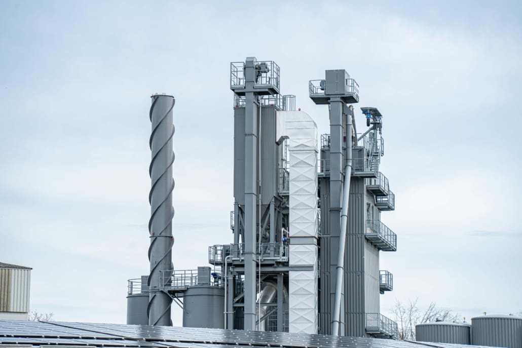 A large industrial facility with tall, gray metal structures, including cylindrical towers and a spiral column, stands against a cloudy sky—ideal for Industrial Asset CGT Valuations. Some buildings and treetops are visible at the bottom of the image.