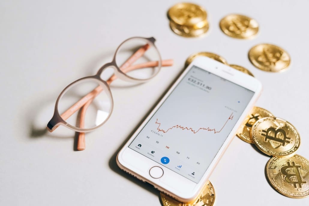 A smartphone displaying a rising cryptocurrency chart, surrounded by physical Bitcoin coins and a pair of eyeglasses on a light surface, illustrating concepts like Goodwill & Intangible Asset CGT Valuations in digital finance.