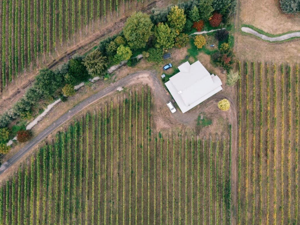 Aerial view of a white house surrounded by trees, nestled between rows of crops or vineyards, with curved dirt roads winding through the landscape—ideal for Rural Property CGT Valuations.