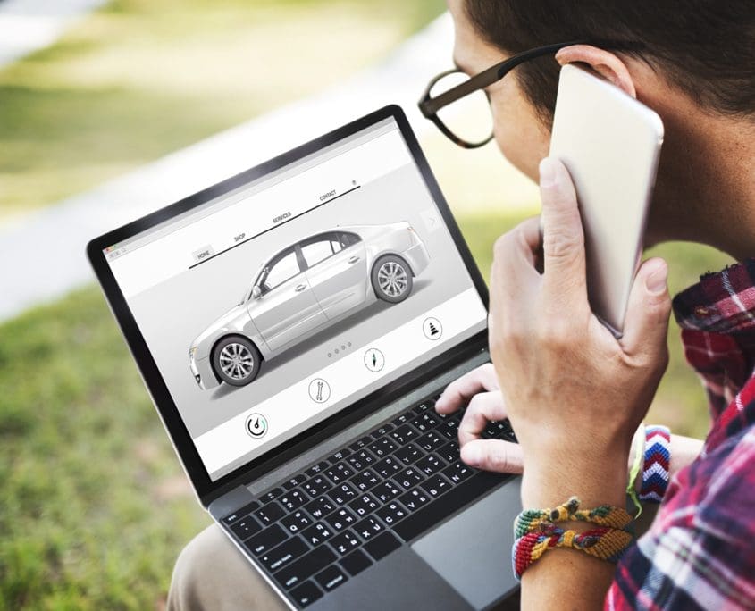 A person wearing glasses and a plaid shirt looks at a laptop showing a digital car model, part of the Asset Valuations Group's latest project. They hold a smartphone to their ear as the screen displays various controls related to the car design, reflecting their focus and expertise.